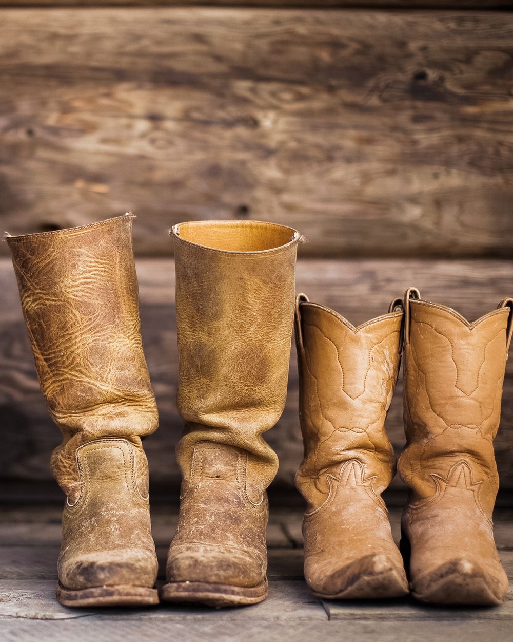 Two worn pairs of tan leather cowboy boots standing on rough planks against a dark log wall